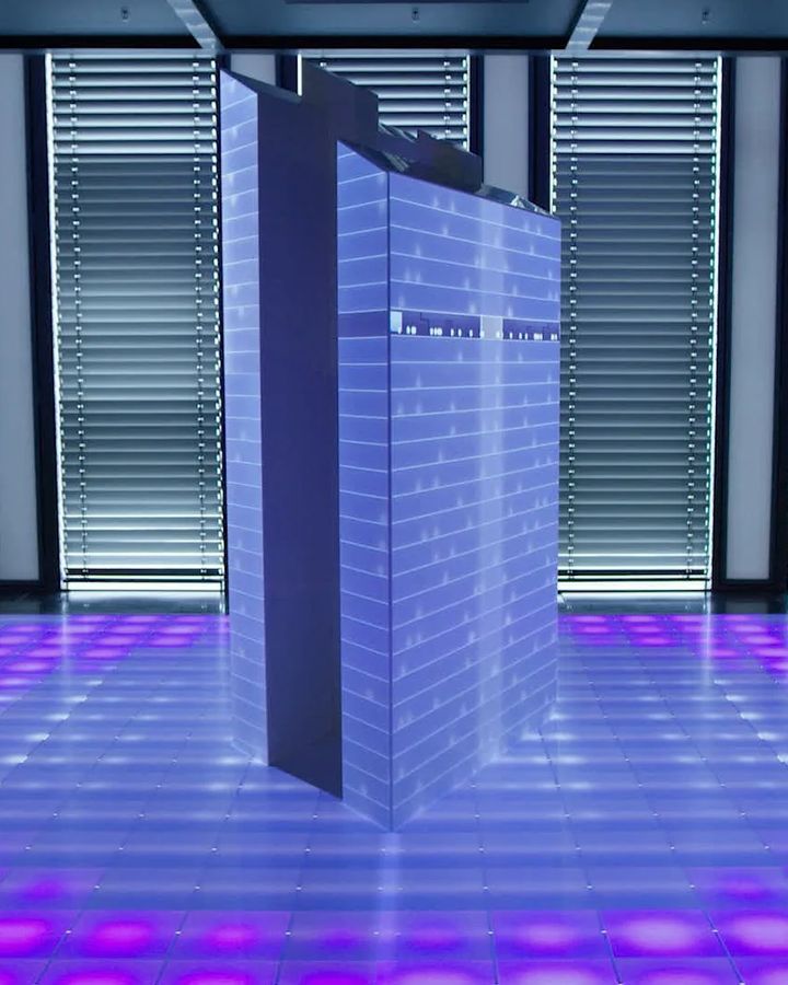 A projection-mapped building model glows above a lit floor grid
